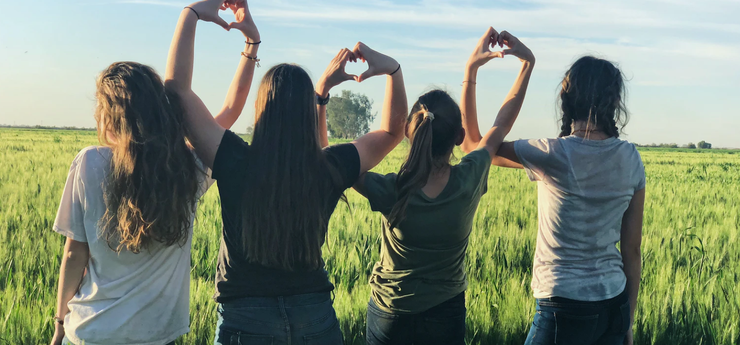 four girls make heart shapes with their hands in a field